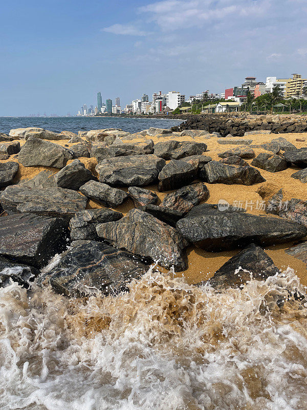 斯里兰卡科伦坡拉维尼亚山海滩的岩石盔甲/撕裂rap和海浪飞溅的图像，沙滩和岩石海滩，背景中可以看到大海和首都，海岸侵蚀防御设施和晴天的蓝天